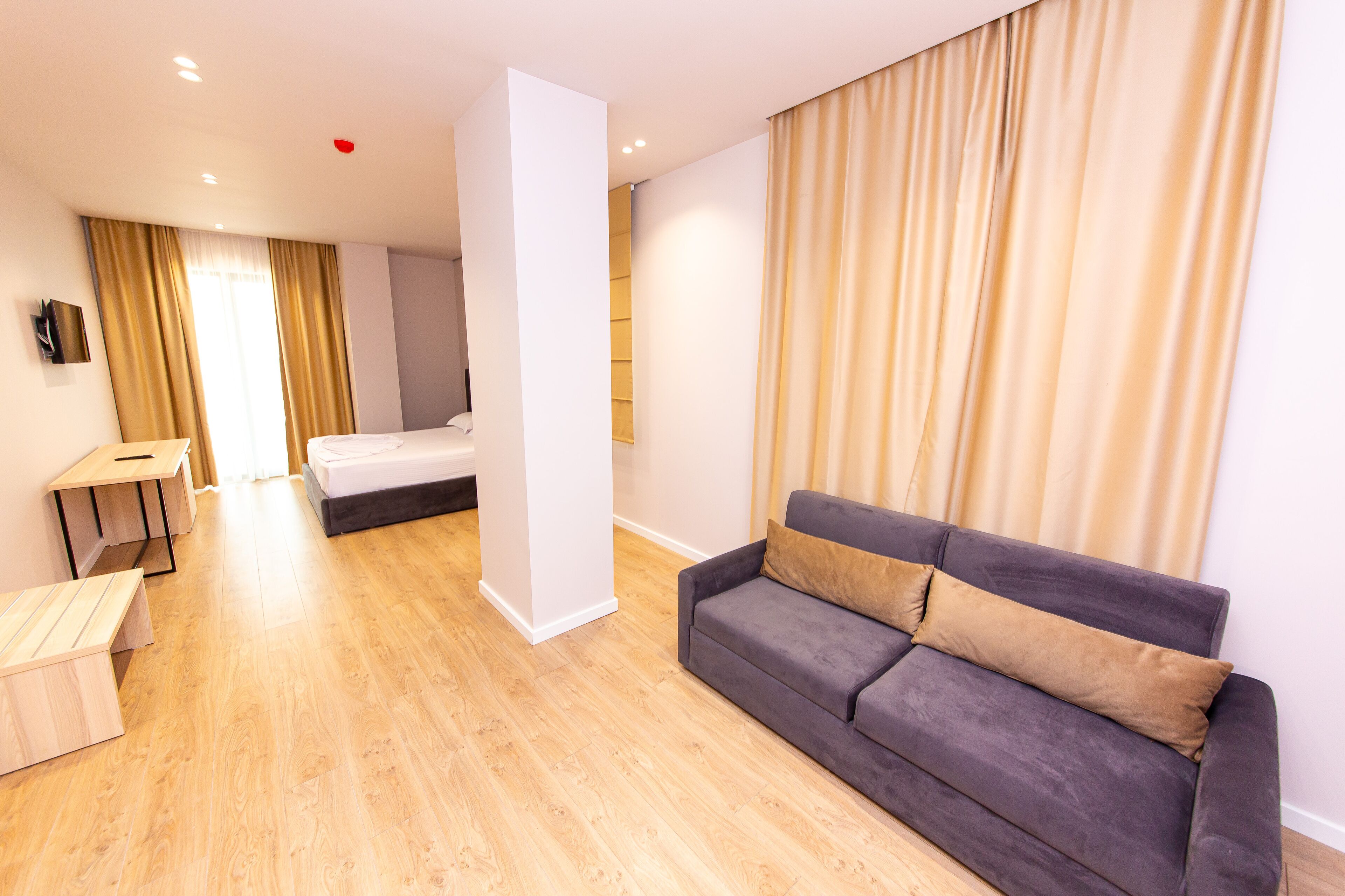 A modern hotel room with a wooden floor, a grey sofa with two cushions, a small desk with a chair, a bed, and a large window with curtains.