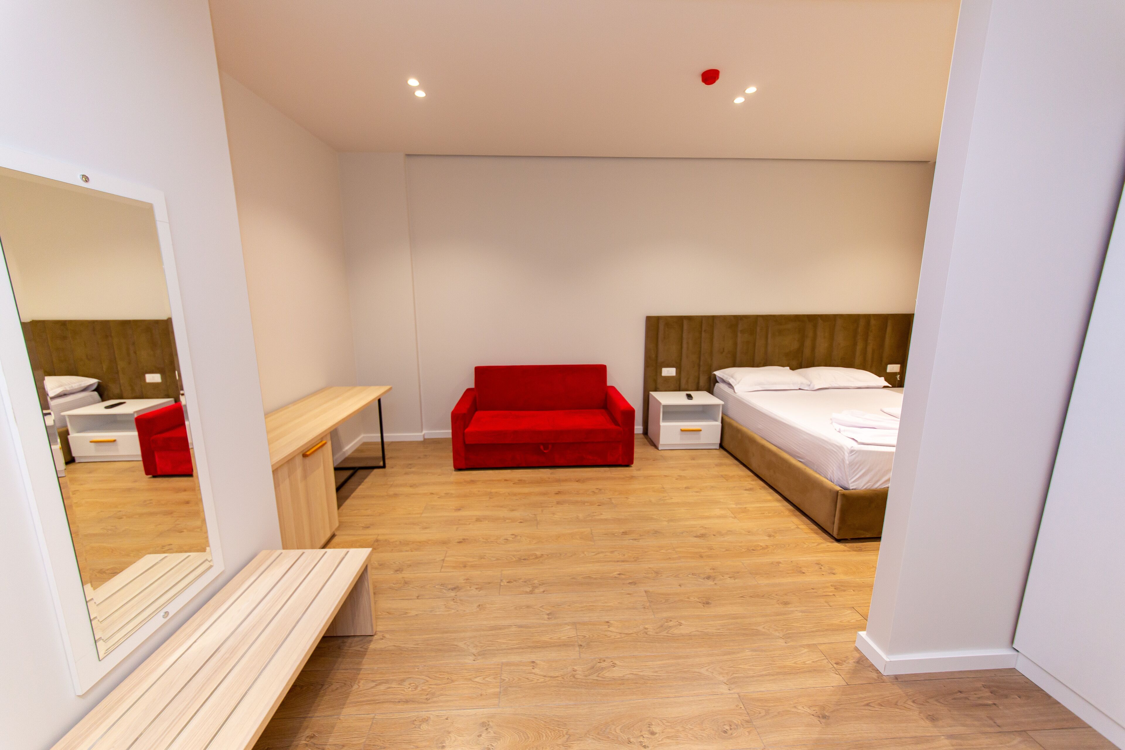 A modern bedroom with a wooden floor, a red sofa, a bed with white bedding, and a nightstand with a lamp.