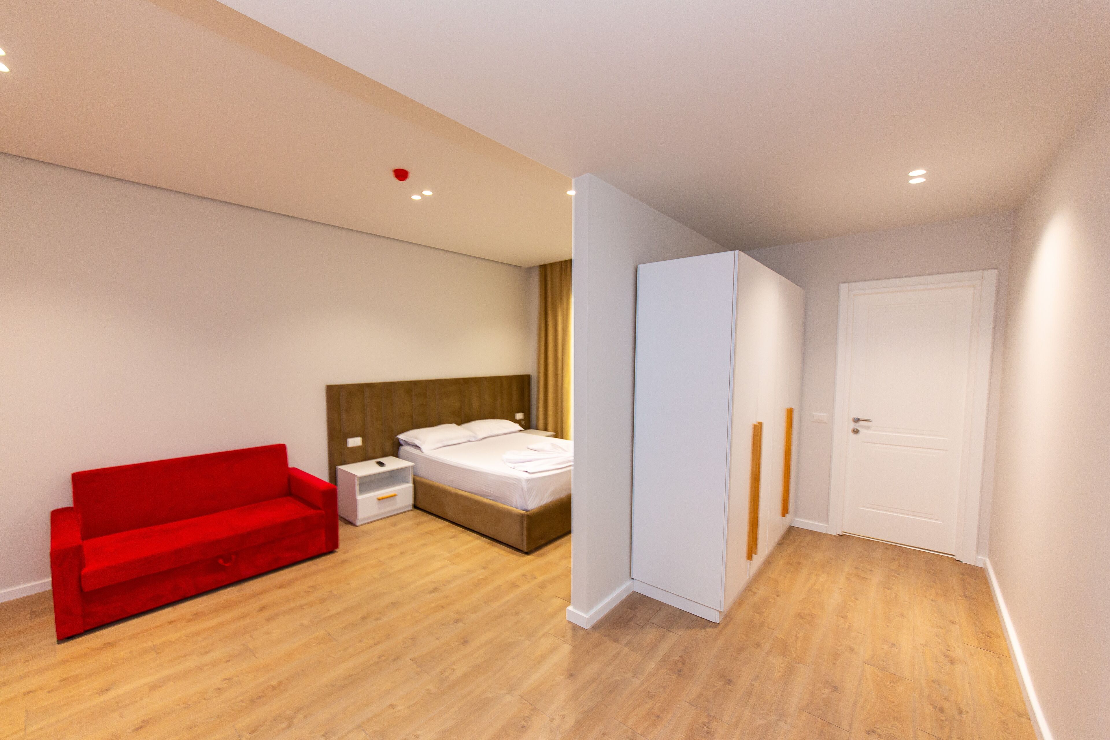 A modern bedroom with a wooden floor, a red sofa, a bed with white bedding, a nightstand, and a wardrobe.