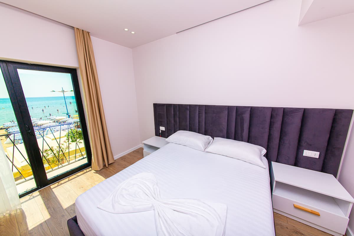 A hotel room with a bed, a nightstand, a bedside table, and a view of the beach and sea through a large window.