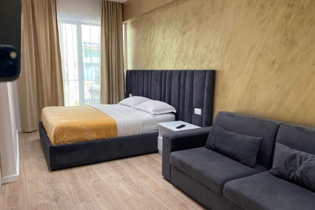 A hotel room with a bed, a sofa, a bedside table, and a window with curtains.