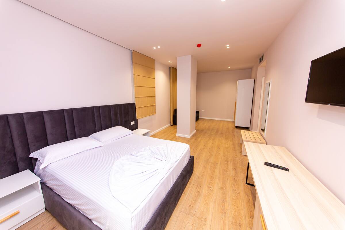 A modern hotel room with a large bed, a bedside table, a desk, and a flat-screen TV mounted on the wall.