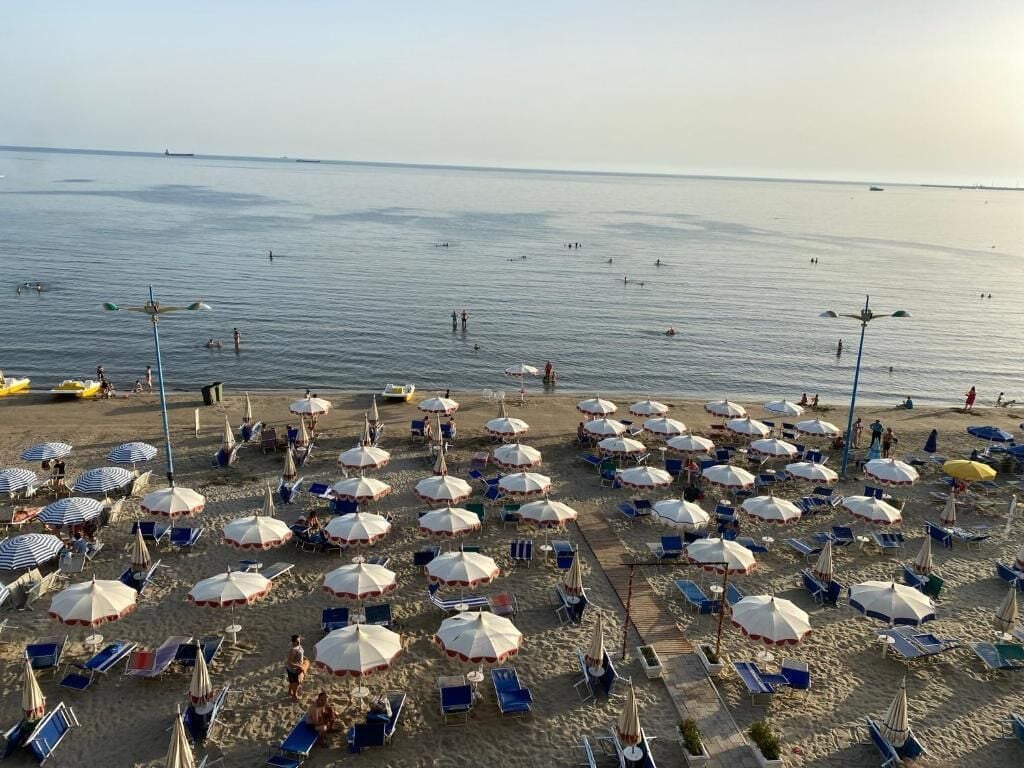 A beach with numerous umbrellas and sun loungers, a calm sea, and a few people in the water.