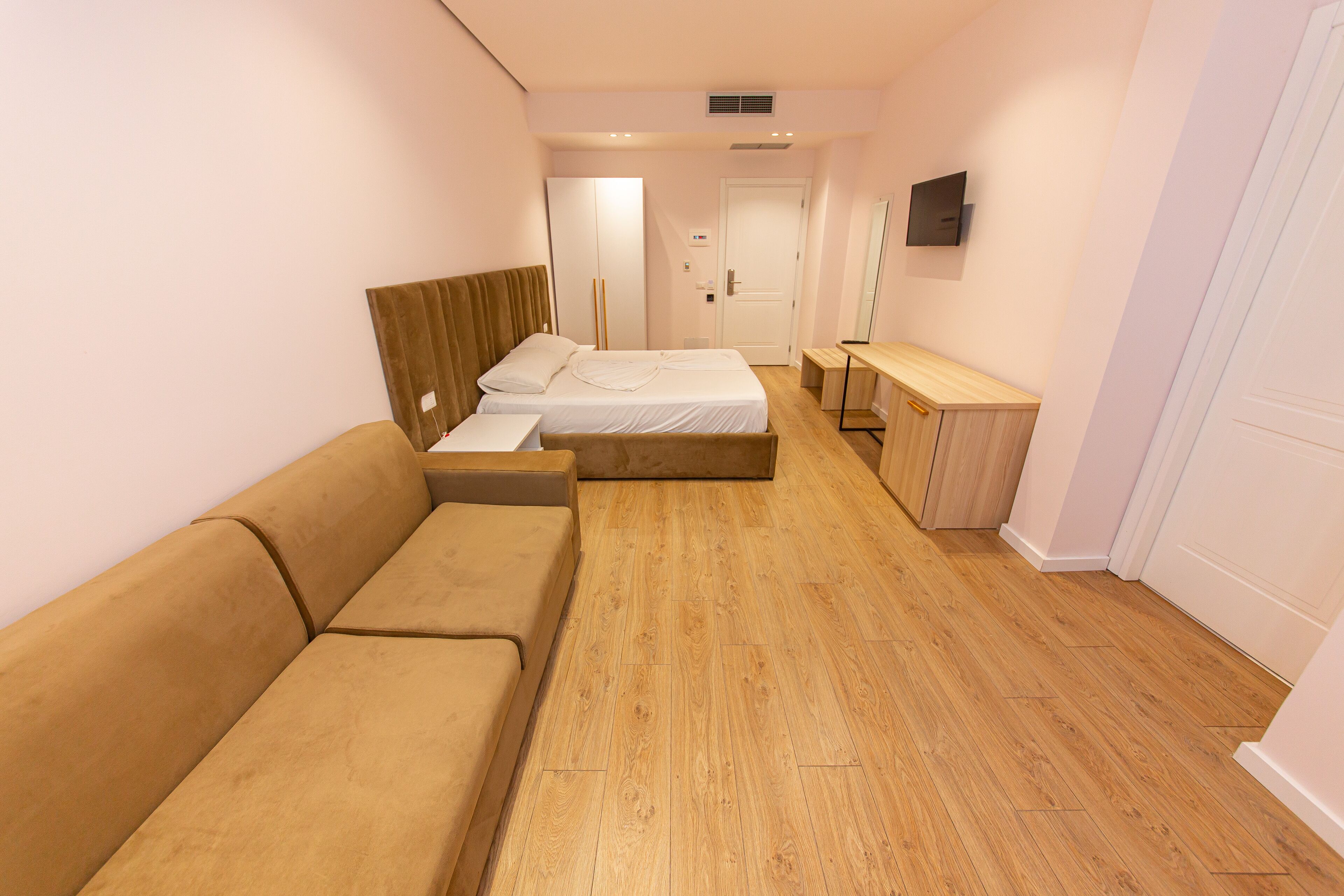 A modern hotel room with a wooden floor, a beige sofa, a bed with white linens, a bedside table, and a mirror on the wall.