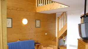 A cozy wooden cabin interior with a blue sofa, wooden dining table, and a staircase leading to an upper level.