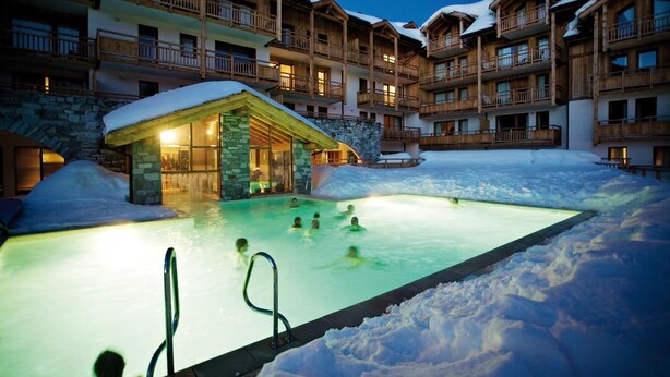 Une piscine extérieure chauffée, entourée de neige, avec des bâtiments en bois en arrière-plan.