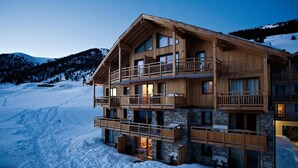 Un chalet en bois à plusieurs étages, doté de balcons, agrémenté de pierres apparentes, et avec une piste de ski en arrière-plan.