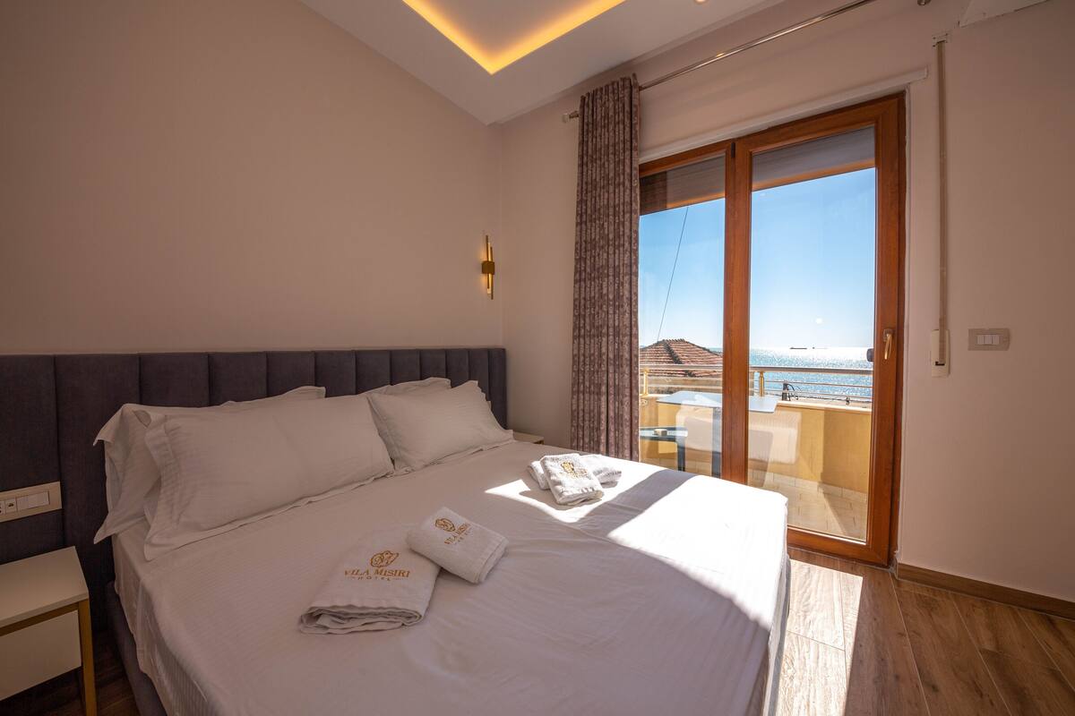 A hotel room with a bed, a nightstand, a large window with a view of the sea, and a sliding glass door to an outdoor area.