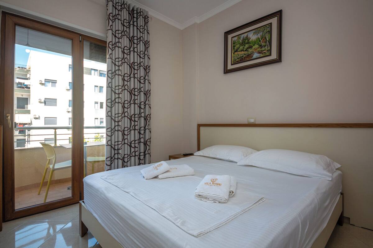 A hotel room with a bed, two pillows, a window with a view of buildings, a curtain, and a painting on the wall.