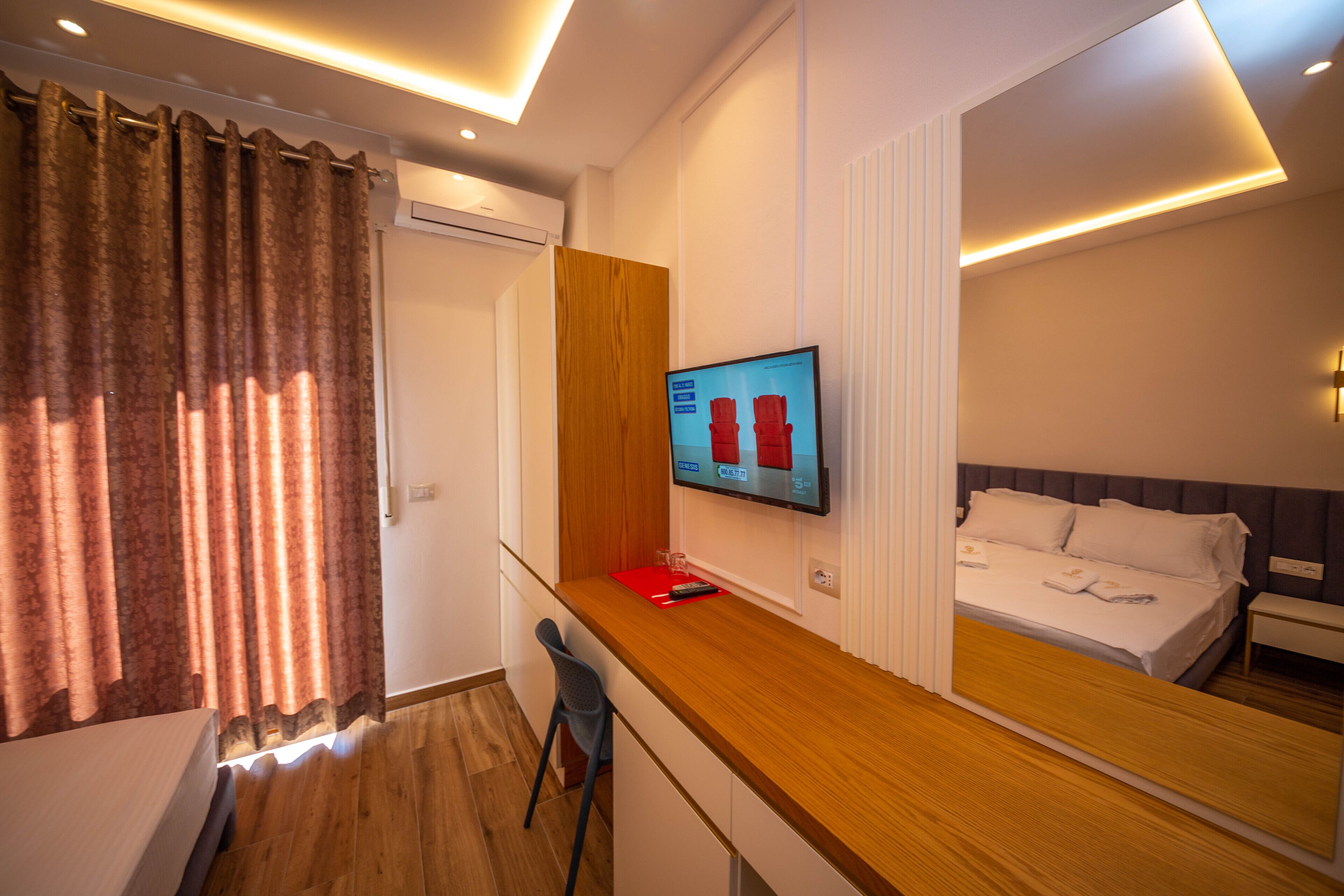 A hotel room with a wooden desk, a TV mounted on the wall, a bed with white linens, and a window with curtains.