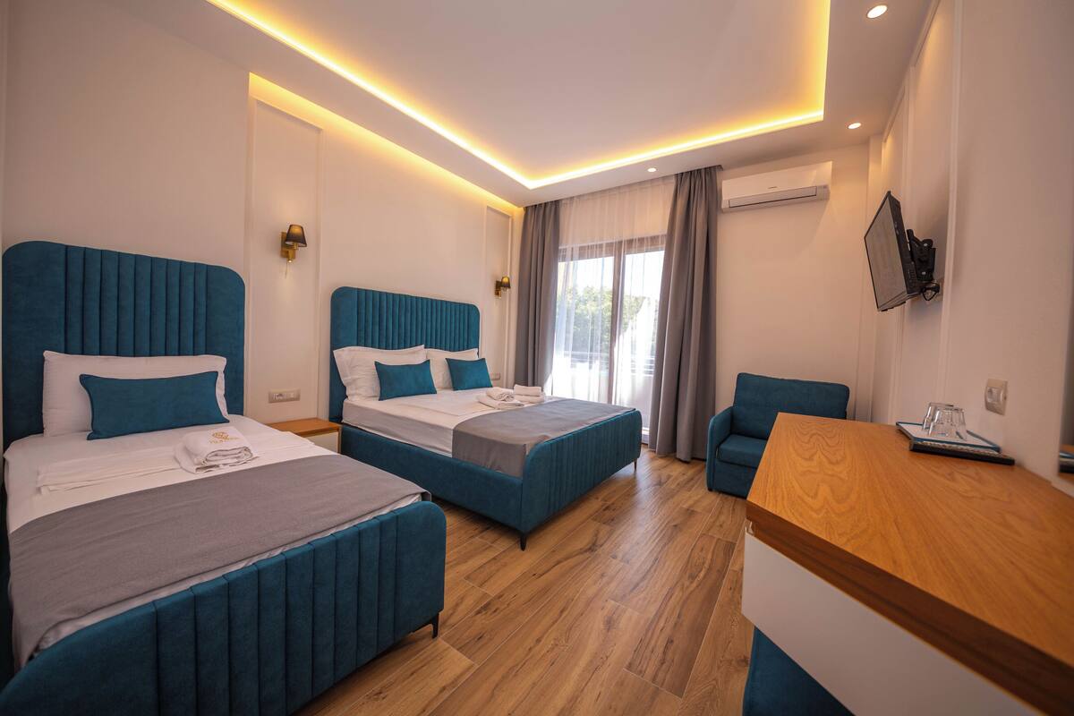 A hotel room with two beds, a wooden desk, a sofa, and a large window with curtains.
