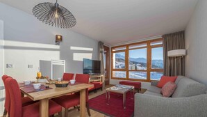 A modern living room with a wooden dining table, red chairs, a grey sofa, and a large window with a mountain view.