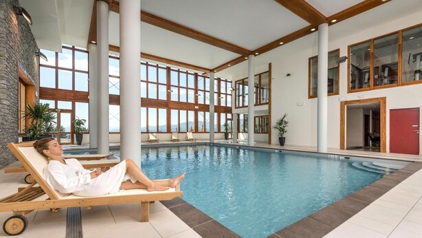 A pool area with a person relaxing on a lounge chair, large windows, and a view of the outdoors.