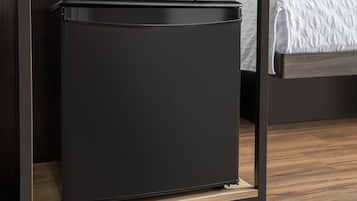 A compact black refrigerator in a modern room with a wooden floor and a bed with a white textured blanket.