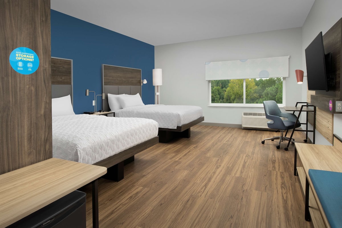 Une chambre d’hôtel avec deux lits, un bureau, une chaise, une télévision et une fenêtre donnant sur des arbres.