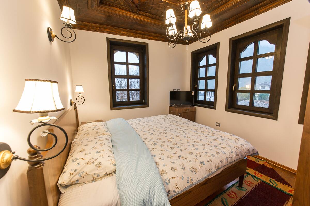 A bedroom with a bed, two windows, a chandelier, a lamp, and a television on a wooden dresser.