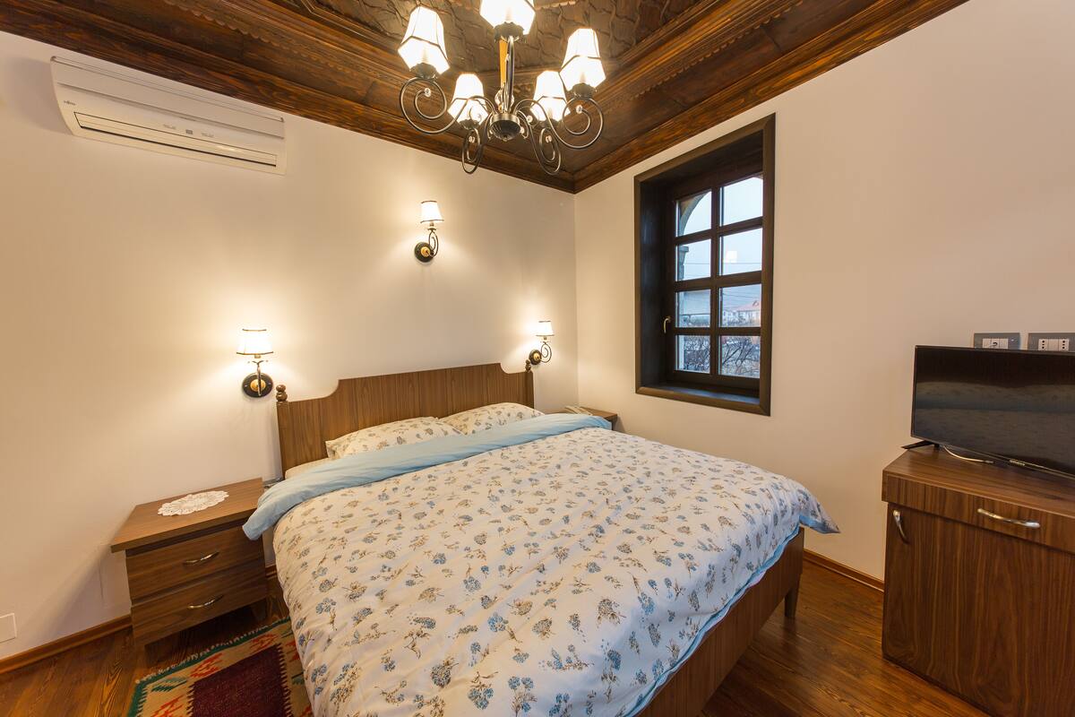 A bedroom with a wooden ceiling, a bed with floral bedding, bedside lamps, a TV on a wooden cabinet, and a window with a view.
