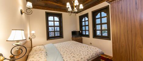 A bedroom with a wooden ceiling, a chandelier, a bed with floral bedding, and two windows.