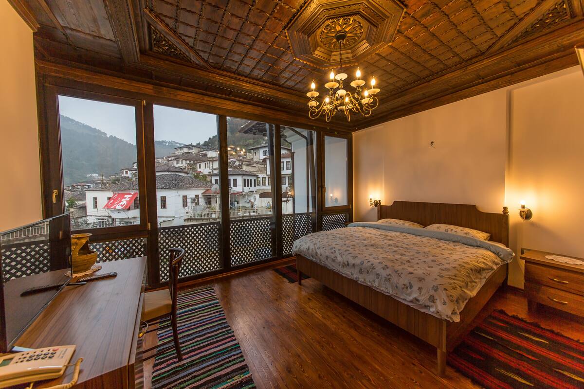 A bedroom with a large window, a wooden desk, a bed with a patterned bedspread, and a chandelier.