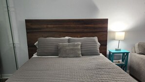 Uma cama com cabeceira de madeira, roupa de cama cinza e uma mesa de cabeceira verde-água com abajur.