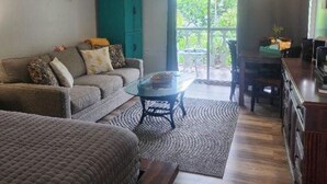 Uma sala de estar acolhedora com sofá, mesa de centro de vidro e ventilador de teto. Uma área de cozinha com mesa e cadeiras visíveis pela porta de vidro deslizante.