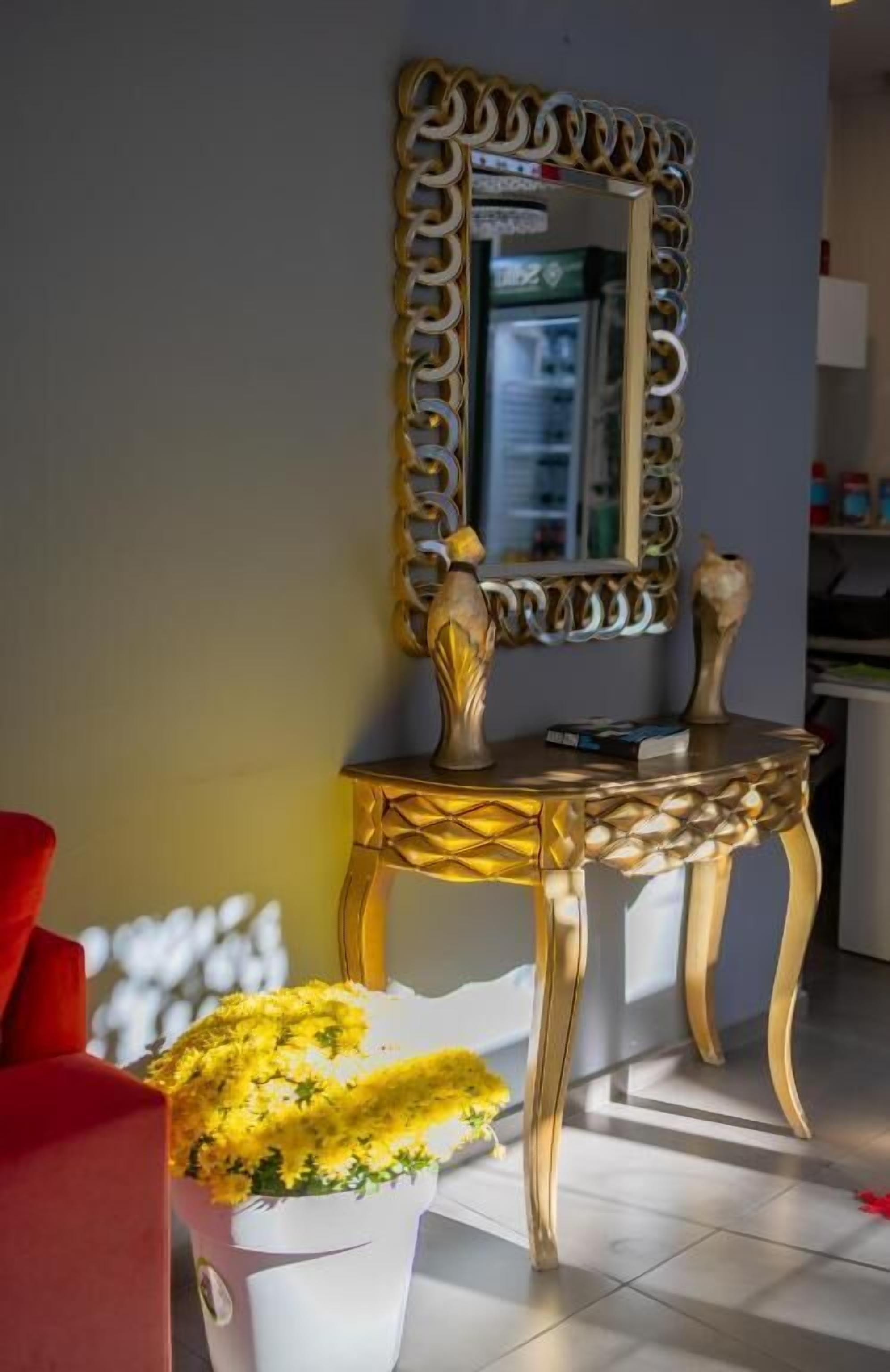 Une pièce avec un miroir encadré d’or, une petite table sur laquelle est posé un vase rempli de fleurs jaunes, et une chaise rouge.