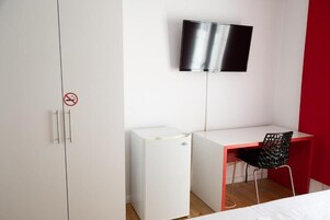 Un dormitorio con un armario blanco, un escritorio blanco, una silla negra, un refrigerador blanco pequeño y un televisor de pantalla plana montado en la pared.
