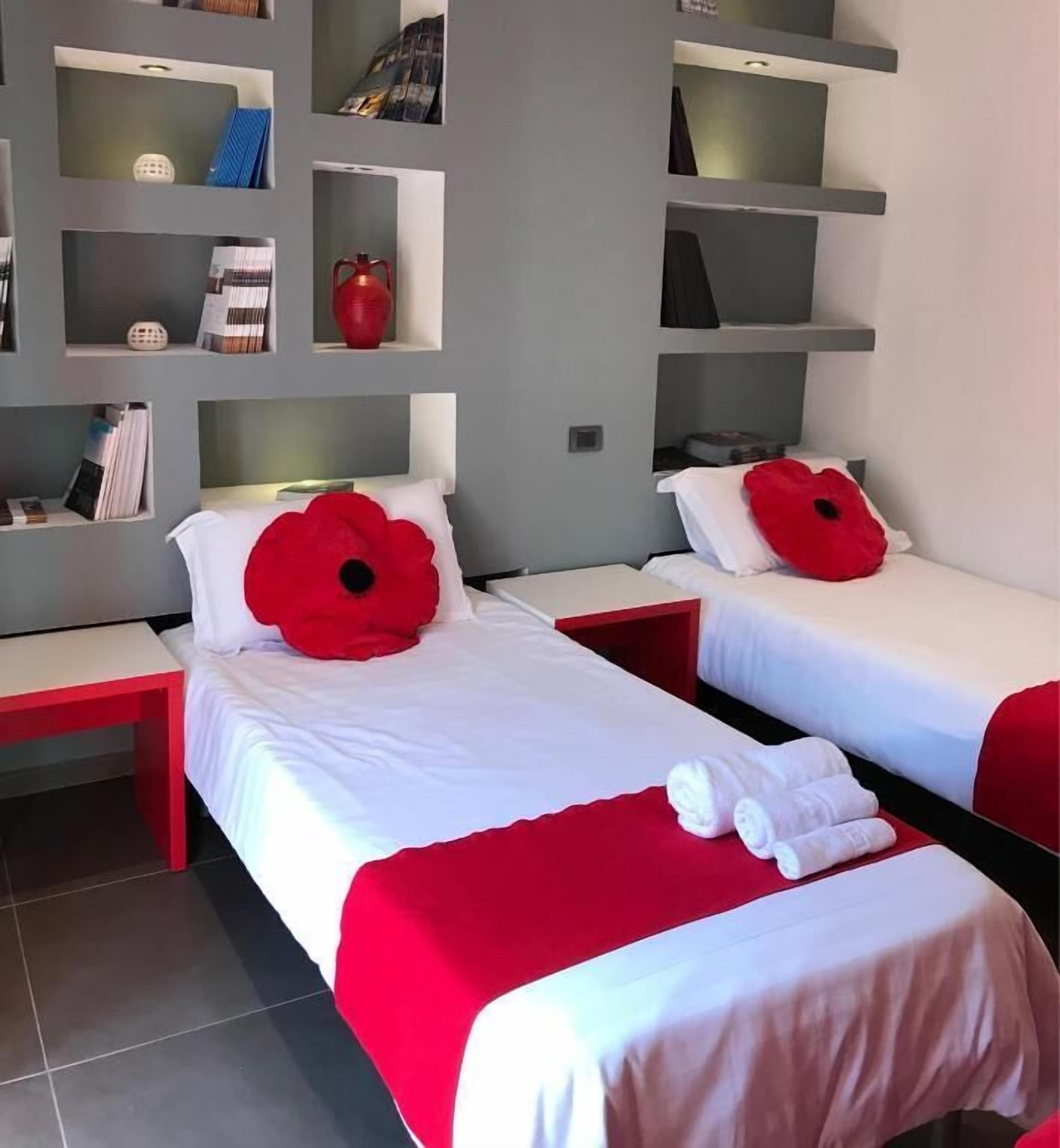 Ein Hotelzimmer mit zwei Betten, jedes mit einem roten Blumenkissen und weißer Bettwäsche. Darüber befinden sich Regale mit verschiedenen Gegenständen.