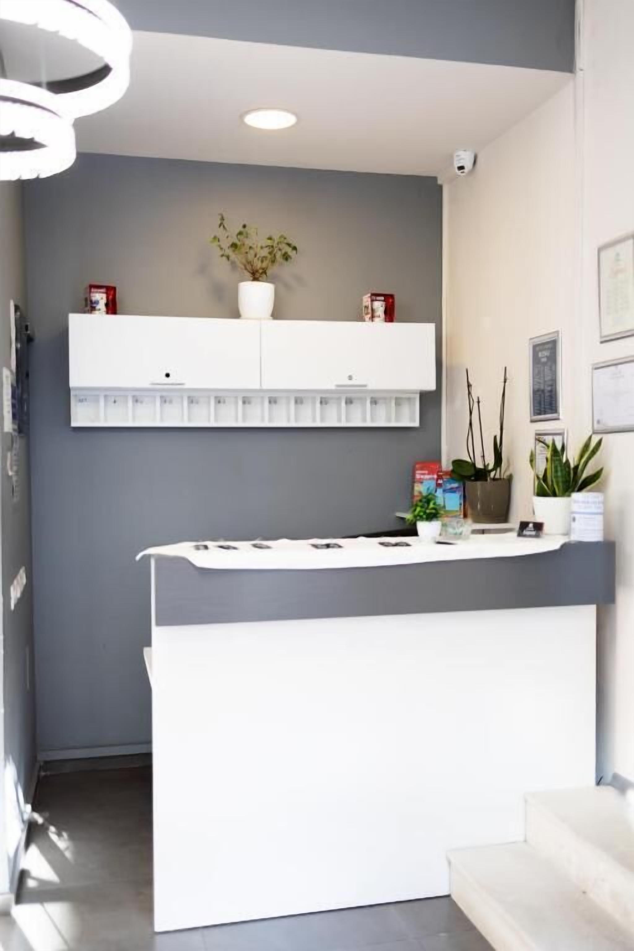 A reception area with a white desk, a potted plant, and a wall-mounted shelf with books and a small plant.
