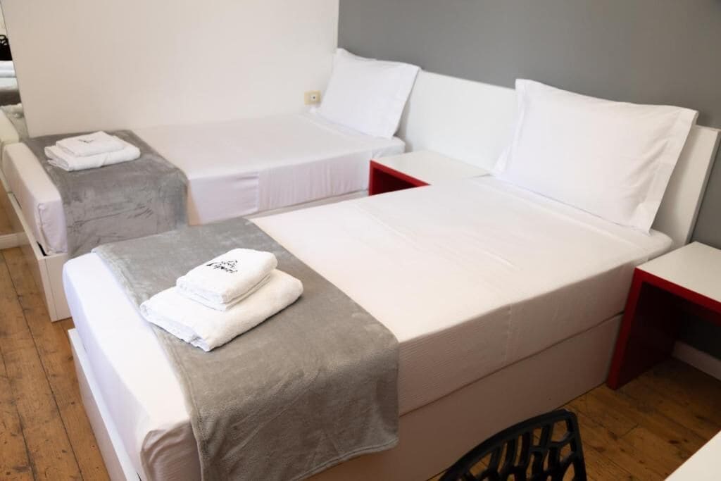 Een hotelkamer met twee eenpersoonsbedden, elk met een witte matras en kussen, en een grijze deken op een van de bedden. Er staat een klein tafeltje met een rode rand en een stoel in de hoek.