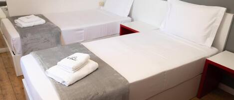 Een hotelkamer met twee eenpersoonsbedden, elk met een witte matras en kussen, en een grijze deken op een van de bedden. Er staat een klein tafeltje met een rode rand en een stoel in de hoek.