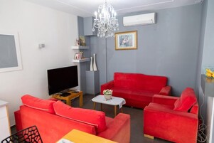 Un salon avec un canapé rouge, un téléviseur à écran plat, un lustre et une petite table sur laquelle est posée une plante.