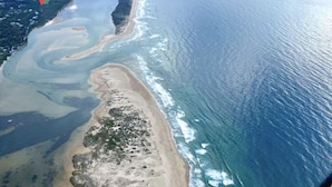 An aerial view of a coastal area with a clear sky, a large body of water, and a narrow strip of land with vegetation.