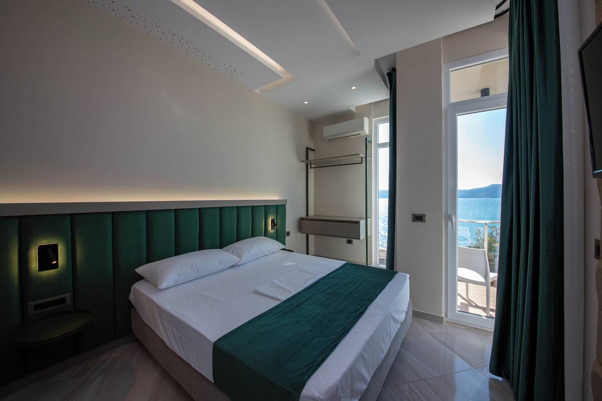A modern hotel room with a large bed, a green accent wall, a built-in TV, and a balcony with a view of the sea.