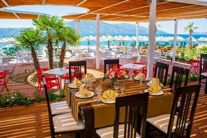 Un área de comedor al aire libre con mesas preparadas para una comida, con vista a una playa y montañas.