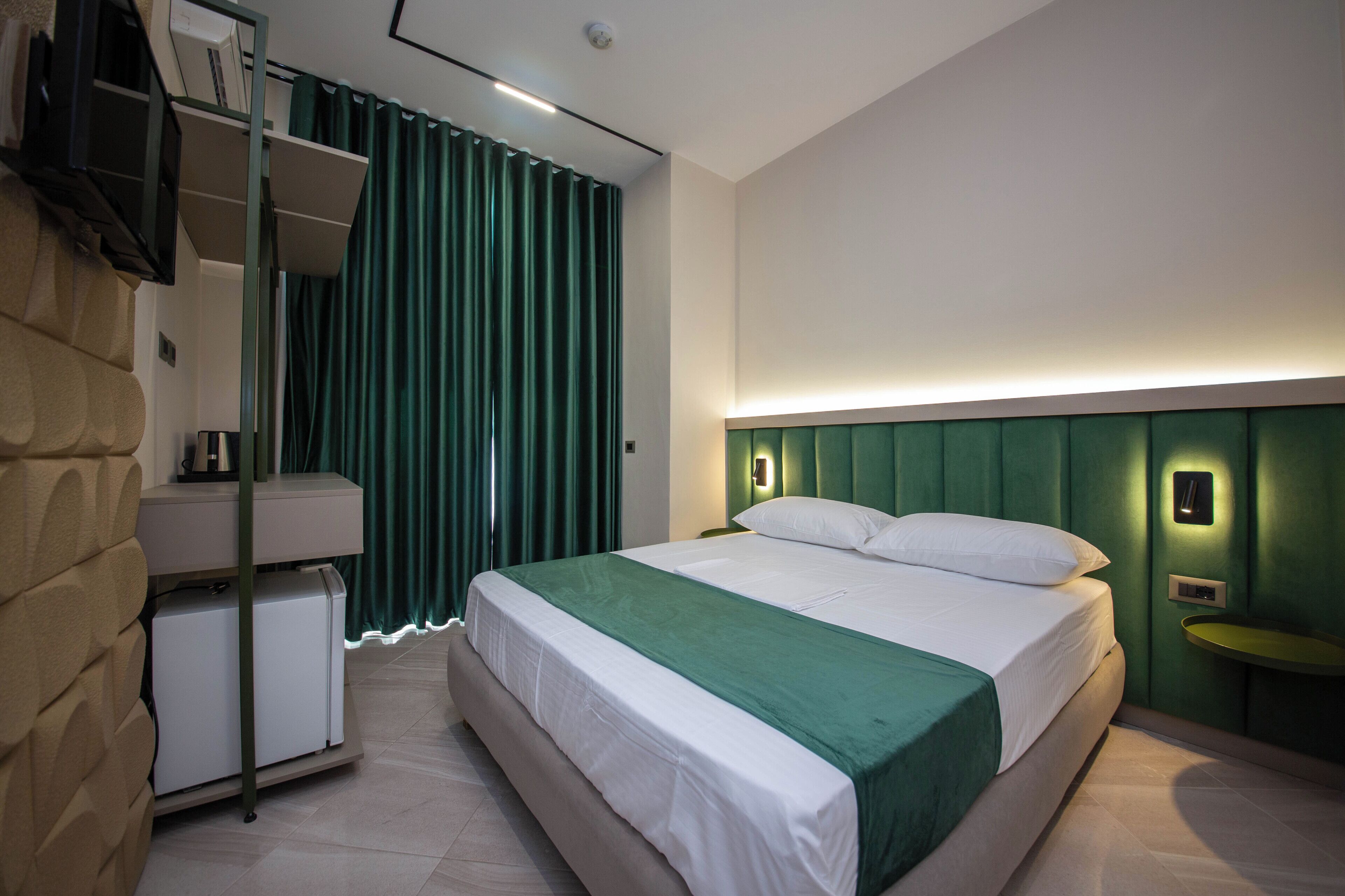 A modern hotel room with a large bed, a bedside table, a built-in shelf, and a green curtain.