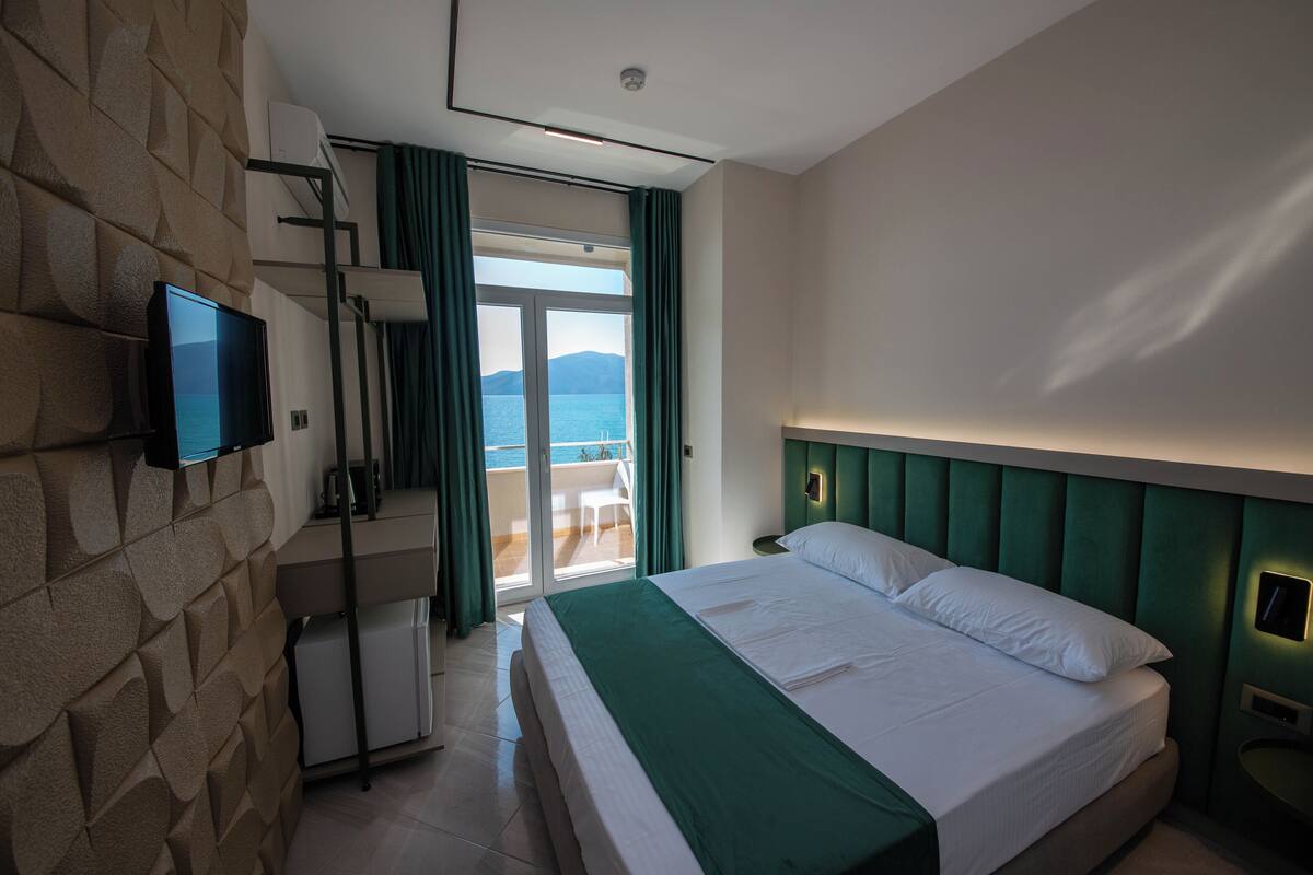 A hotel room with a bed, a television on the wall, a balcony with a view of the sea, and a green headboard.
