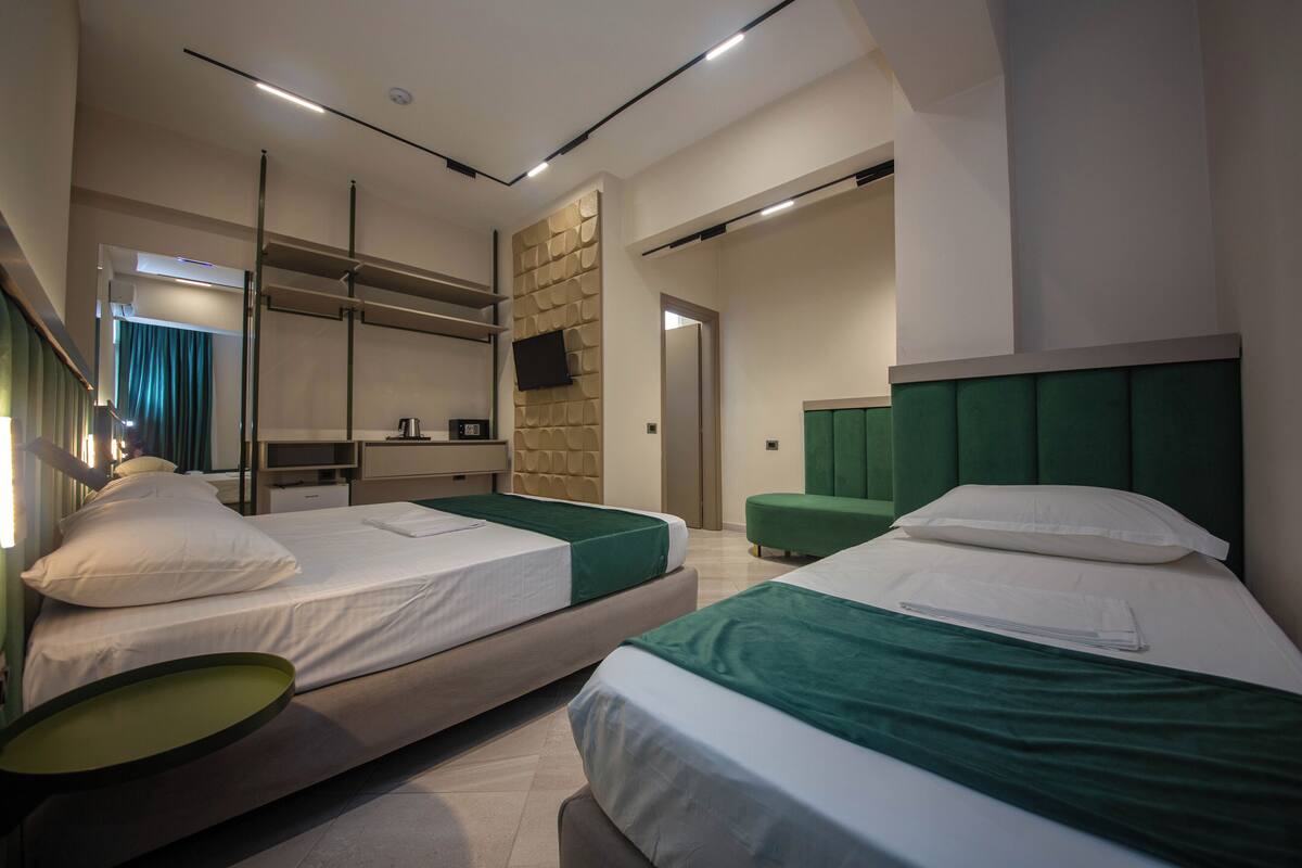 A hotel room with two beds, a green accent wall, a flat-screen TV mounted on the wall, and a small kitchenette area.