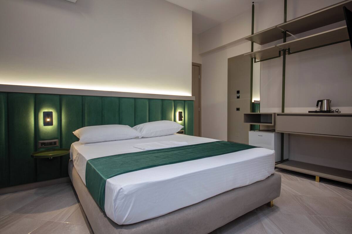 A modern hotel room with a large bed, a bedside table, a mirror, and a built-in wardrobe.