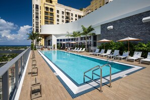 Un área de piscina en la azotea con sillones y sombrillas, rodeada por un edificio moderno con balcones.