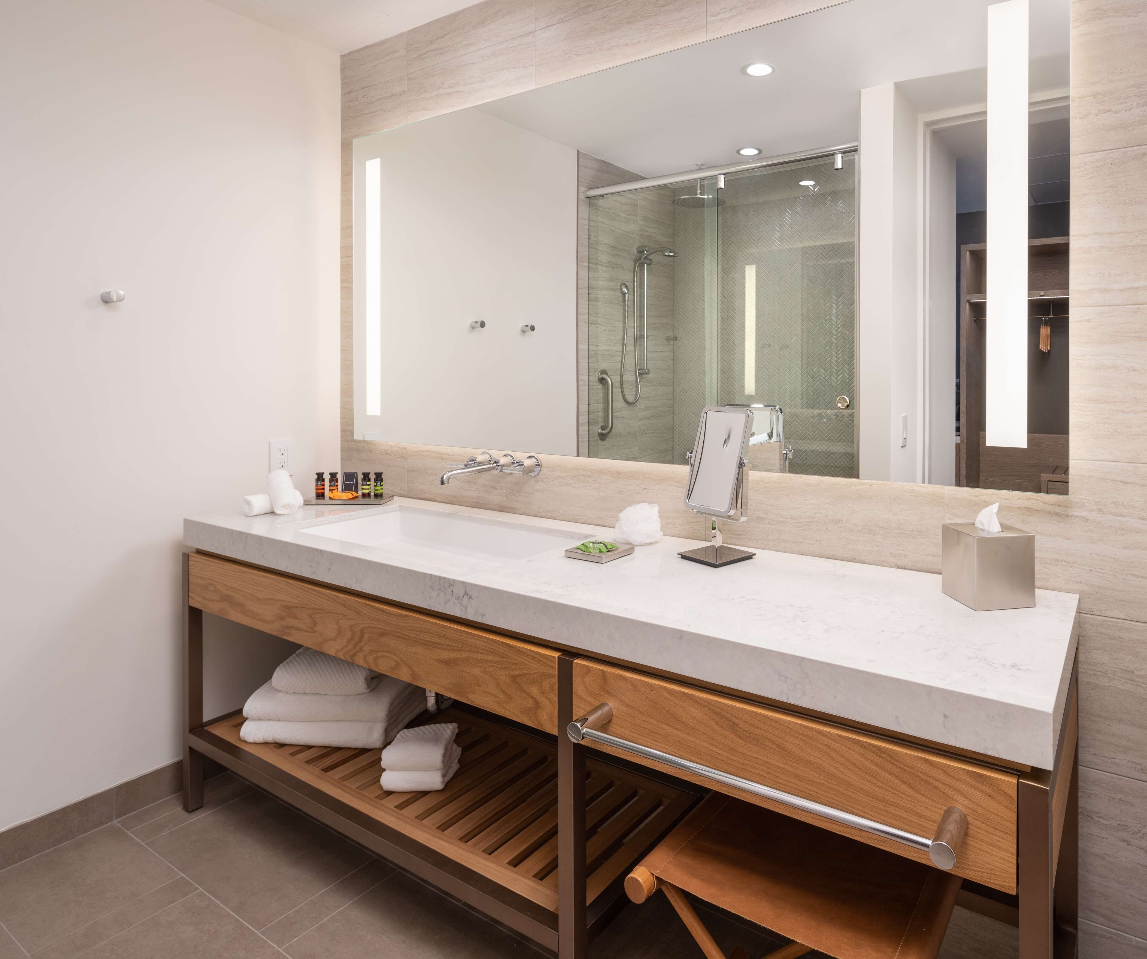 A modern bathroom with a large mirror, two sinks, a shower, and a wooden vanity with storage.