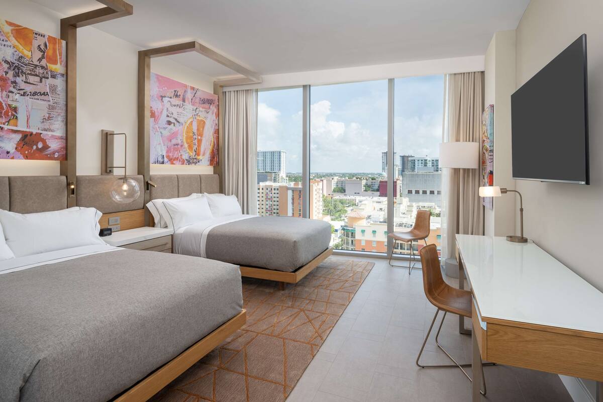 Ein Hotelzimmer mit zwei Betten, einem Schreibtisch und einem großen Fenster mit Blick auf die Stadt.