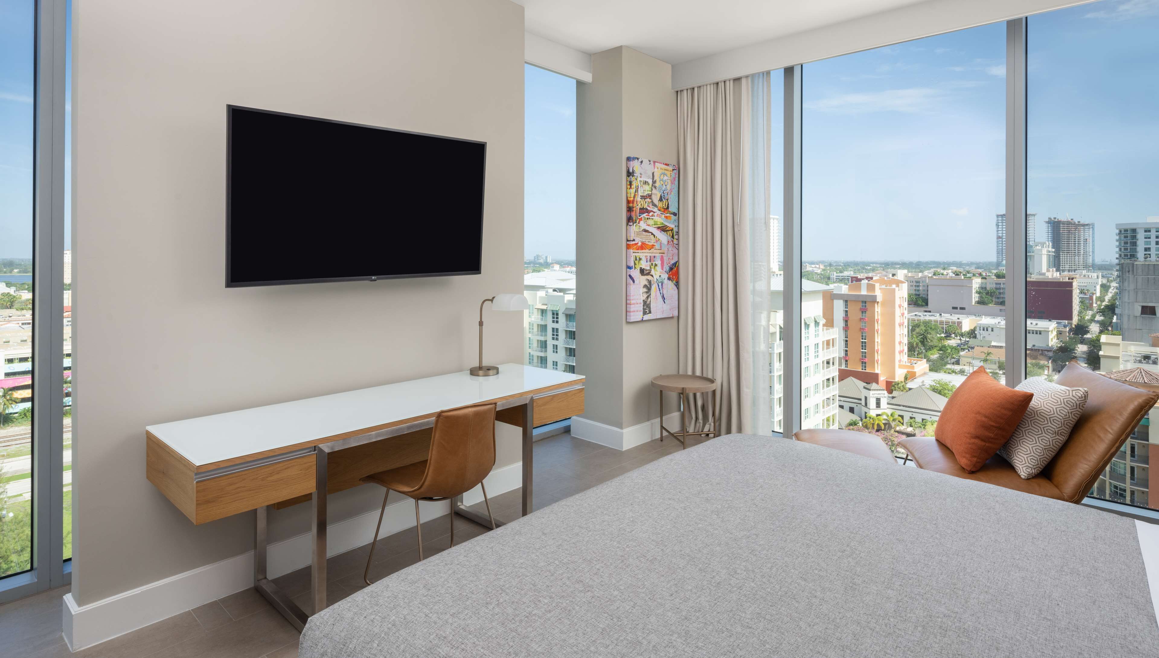 A modern hotel room with a large bed, a desk with a chair, a television, and a large window offering a city view.