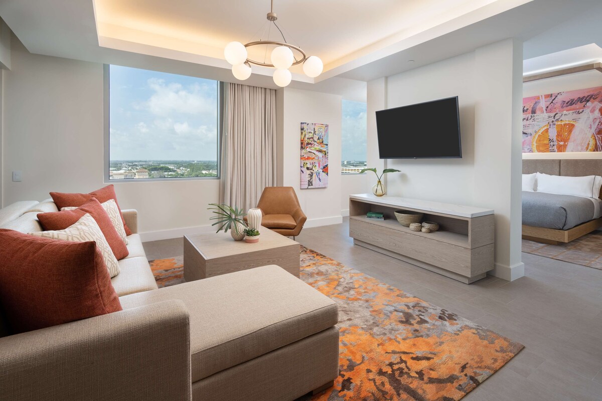 A modern living room with a sofa, armchair, and coffee table, a flat-screen TV mounted on the wall, and a large window offering a city view.