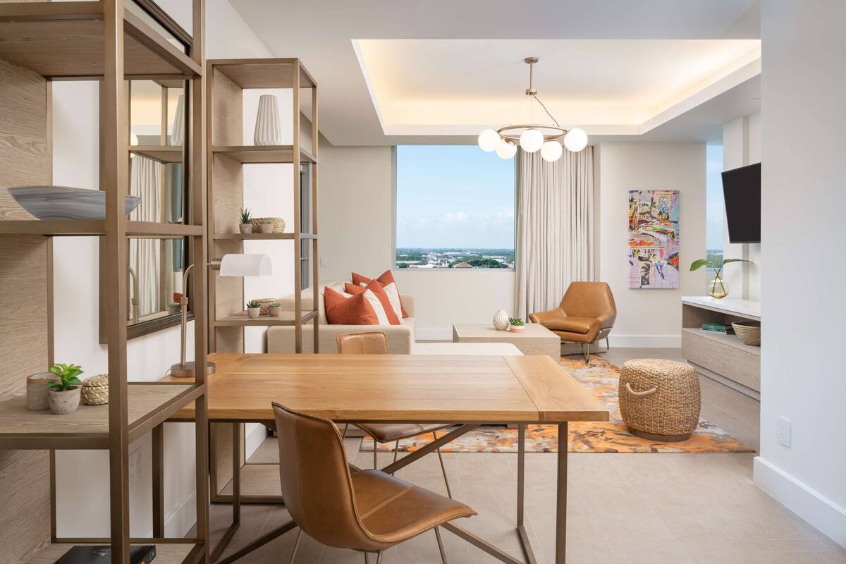 Ein modernes Wohnzimmer mit einem Holzesstisch, Ledersesseln, einem Sofa mit orangenen Kissen, einem Fernseher und einem großen Fenster mit Blick auf die Stadt.