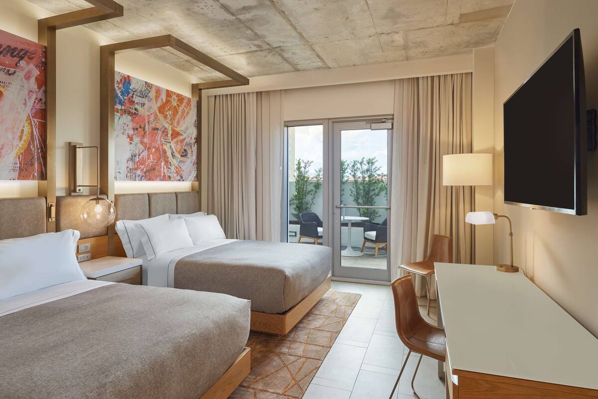 Ein Hotelzimmer mit zwei Betten, einem Schreibtisch, einem Fernseher und einem Balkon mit Blick auf Bäume und Gebäude.