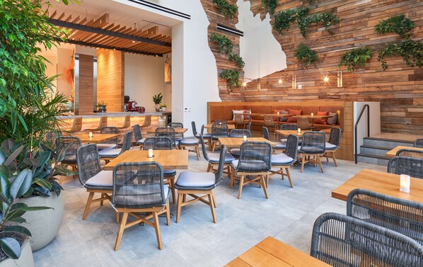 Un café moderno con mesas y sillas de madera, una zona de bar y plantas verdes en las paredes.