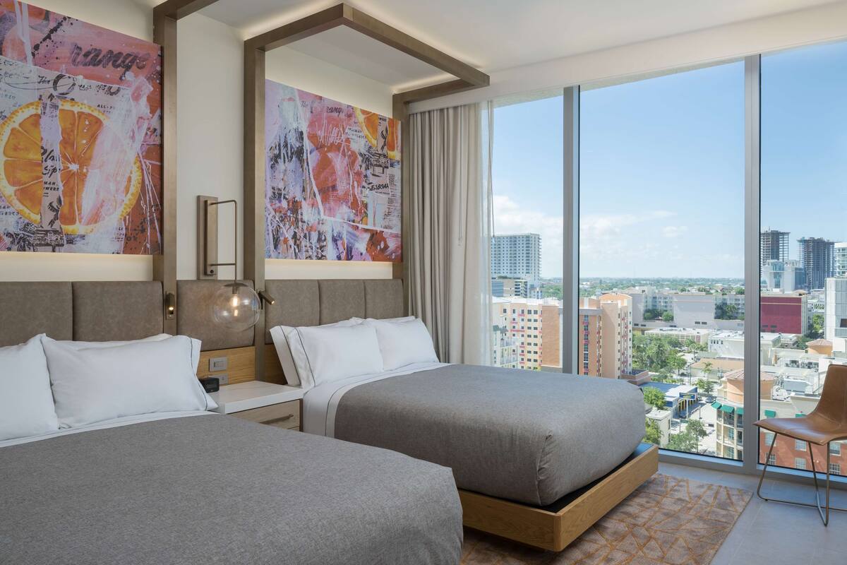 Ein Hotelzimmer mit zwei Betten, einem großen Fenster mit Blick auf die Stadt und Kunstwerken an den Wänden.