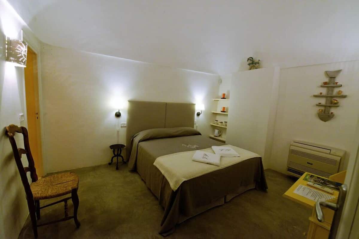 A small, cozy bedroom with a single bed, a chair, a bedside table with a lamp, a shelf with toiletries, and a radiator.