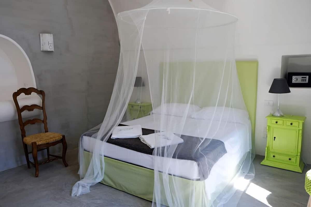 A bedroom with a bed under a mosquito net, a wooden chair, a green nightstand with a lamp, and a wall-mounted light fixture.